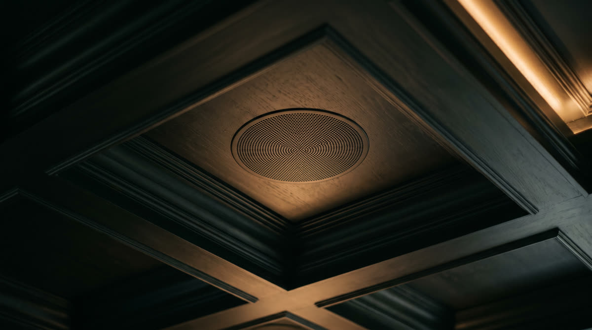 Close-up of an in-ceiling round speaker grille flush-mounted in a dark coffered ceiling with warm amber accent light